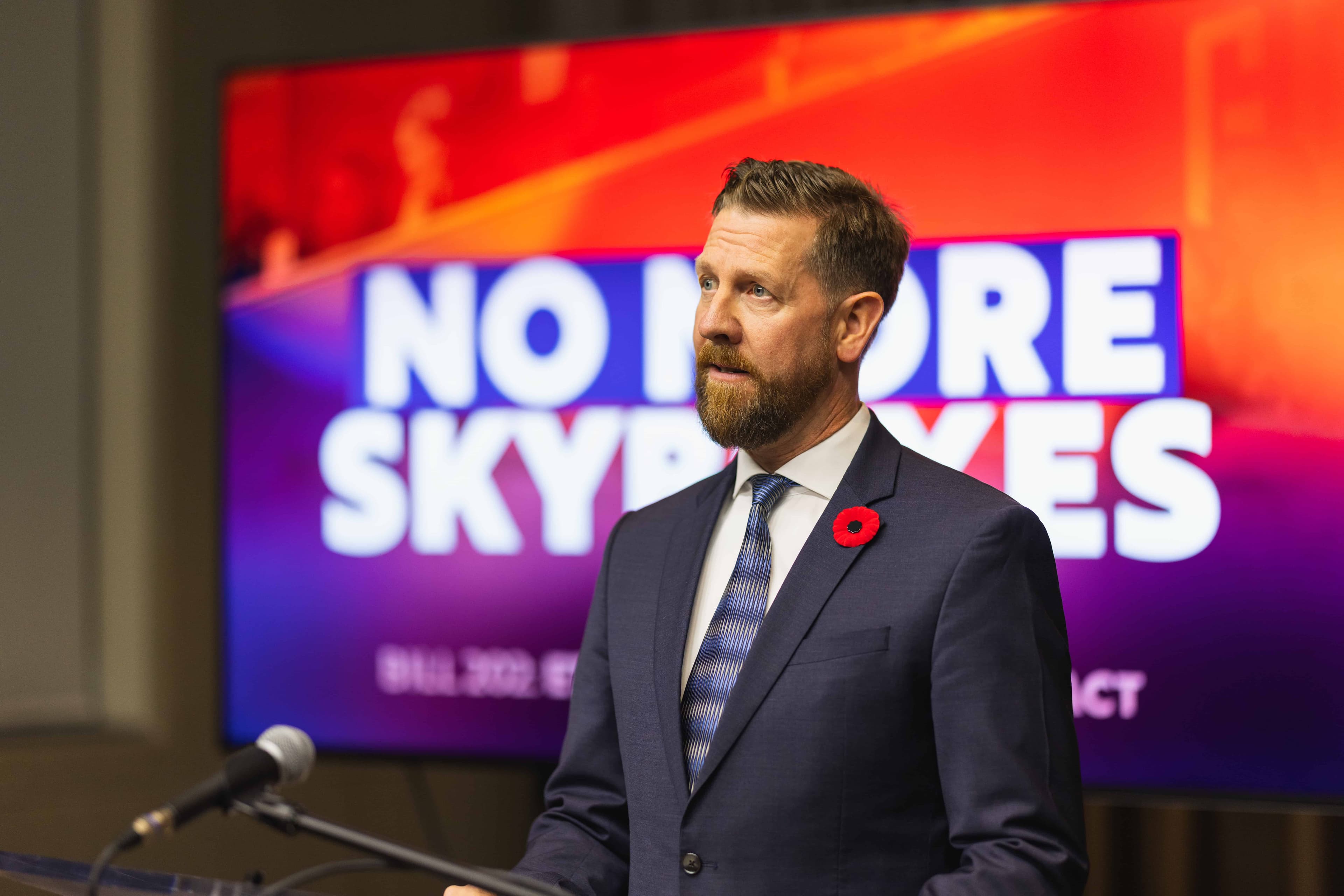 MLA Kasawski speaks at a press conference, standing in front of a screen reading "No More Skyboxes"