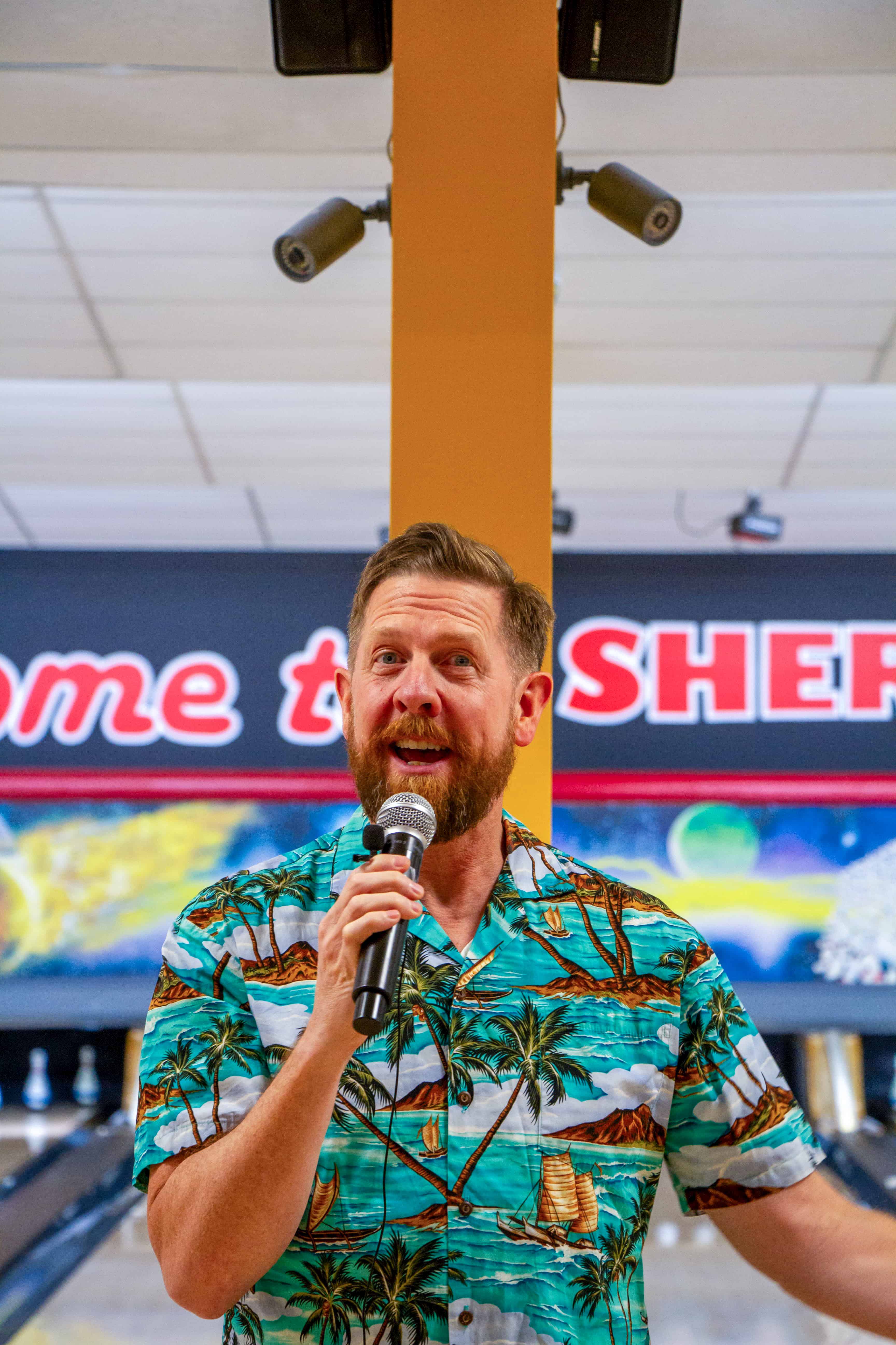 Kyle wearing Hawaiian theme shirt holding a microphone at the bowling alley.